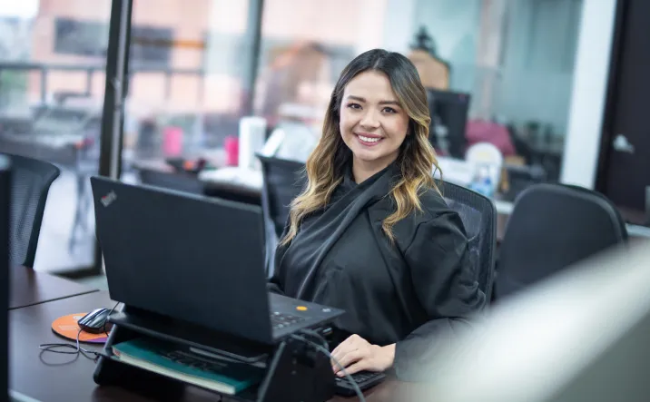Mujer sonriendo sentada en una silla. Frente a ella un escritorio con un computador portátil.